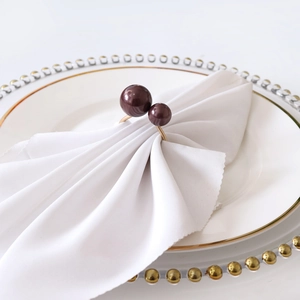 Detalle de servilletero dorado con cuentas burdeos sobre mantel blanco y plato