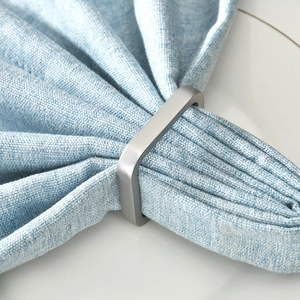 Detail of square matte silver napkin ring on blue linen fabric with brushed metal finish