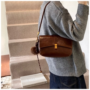 Detail of a dark brown leather crossbody bag with a gold buckle and plush pom-pom charm