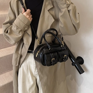 Glossy black bowling handbag with gold hardware and front flap pockets worn with a trench coat