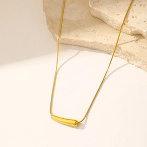 Pendentif barre dorée polie sur une chaîne serpent posée sur une pierre beige