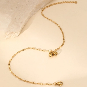 Cavigliera dorata con catena a foglia e campanellini su sfondo beige dallo stile minimalista