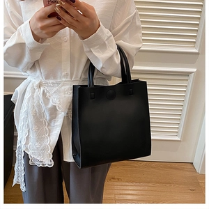 Vertical black tote bag with dual handles and round metal buckle detail in a room setting