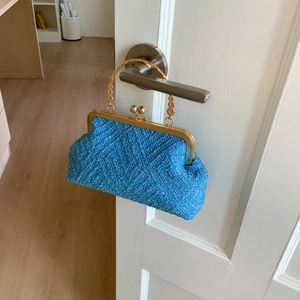 Blue woven shell bag with gold metal handle hanging on a door handle in a home setting