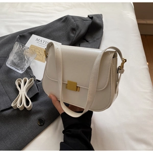 Cream white rectangular crossbody bag with gold buckle and two shoulder straps on a bed