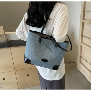 Rear view of a large denim tote bag with black leather trim worn by a model in a minimalist room