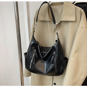 Detail van zwarte glanzende hobo-tas met plooistructuur en ritsen op een beige jas
