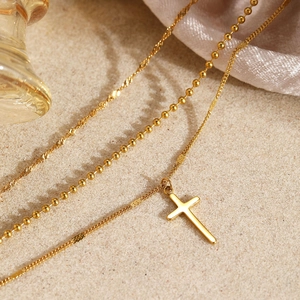 Collier multi-rangs avec pendentif croix dorée sur un fond de texture sablonneuse
