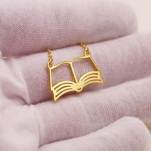 Detail of a hollow book pendant in gold stainless steel held by a gloved hand