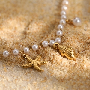 Detail of seashell and starfish charms on a pearl anklet against a sandy beach backdrop