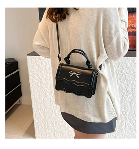 Detail of black crossbody bag with golden bow clasp and woven handle on white knit background