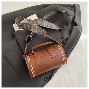 Detailed view of brown cylindrical crossbody bag with gold rivets and patterned wide strap