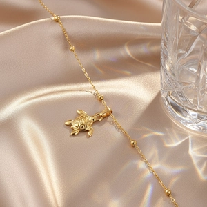 Minimalist stainless steel turtle pendant anklet with golden beads on satin background
