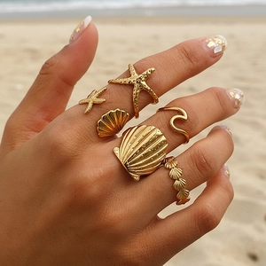 Close-up of a 6-piece gold starfish and shell ring set worn on a summer beach background