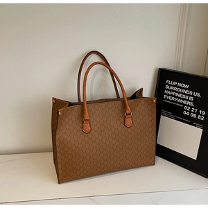Detail of caramel tote bag with M-pattern print, metal rivets, and orange handles