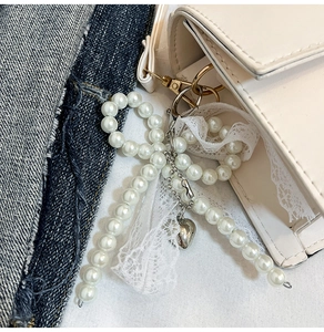 Detail of pearl bow charm with lace trim and silver heart pendant on a beige handbag