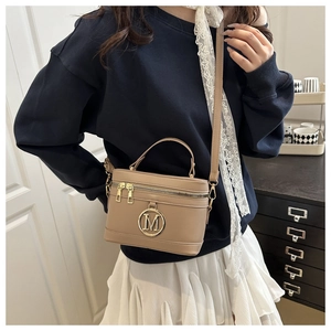 Detail of a beige box-shaped crossbody bag with a metal M logo on a female model