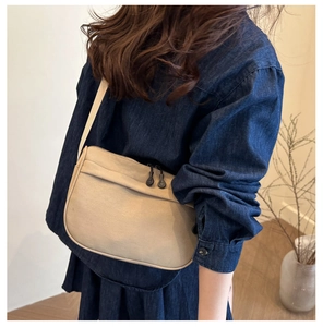 Beige crescent crossbody bag with wide strap worn over denim jacket in soft home setting