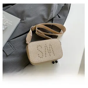 Detail of beige woven crossbody bag with silver SM rivet logo and wide strap next to phone
