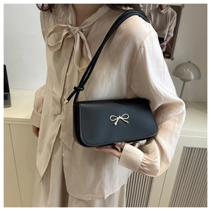 Detail of black glossy shoulder bag with golden bow buckle on model in beige shirt
