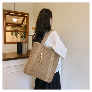 Detail of a large woven straw tote bag with tassel charm worn by a model in a cafe