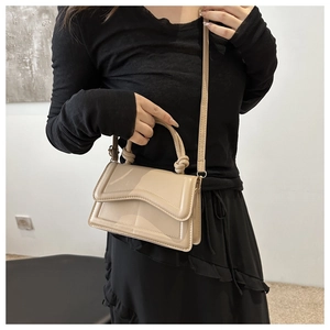 Detail van beige glanzende vierkante handtas met golvende flap en knoop op het handvat