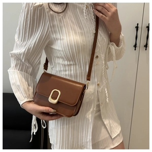 Brown faux leather crossbody bag with golden oval buckle worn by model in white dress