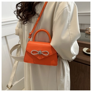 Orange leather handbag with rhinestone bow detail shown worn over a white coat