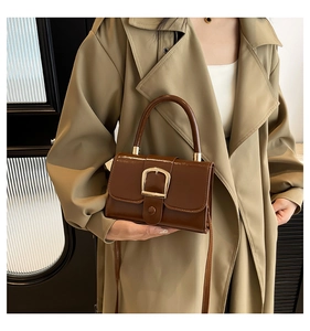 Detail of a brown patent leather crossbody bag with a gold buckle on a khaki trench coat