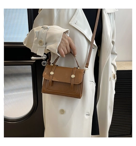 Detail der braunen Umhängetasche mit Goldschnallen an einem eleganten weißen Trenchcoat