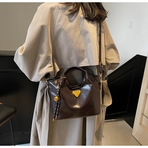 Détail du sac seau en cuir verni marron foncé avec logo triangle doré sur un trench-coat beige