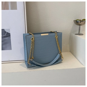 Close-up of light blue crocodile texture handbag with gold chain straps on white podium