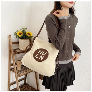 Detail of a beige canvas shoulder bag with brown round logo and slim straps on a model