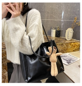 Detail of a black high-gloss tote bag with a beige plush bear charm on white sweater