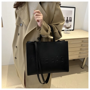 Detail of black leather tote bag with embossed TOTE BAG text carried by model in trench coat