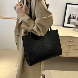 Black leather large tote bag with embossed letters worn by a model in a trench coat