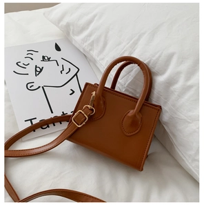 Brown mini crossbody bag with gold hardware placed on a white pillow in a minimalist setting