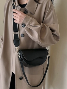 Matte black saddle bag with wavy flap and striped wide strap shown on a camel trench coat