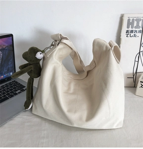 Close-up of beige canvas shoulder bag featuring a green plush frog charm and wide strap