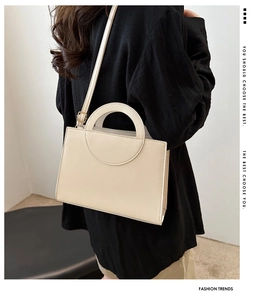 Model wearing a structured cream crossbody bag with a circular handle and gold hardware