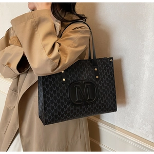 Schwarze Monogramm-Einkaufstasche mit geprägtem M-Logo, am Model mit Trenchcoat gezeigt