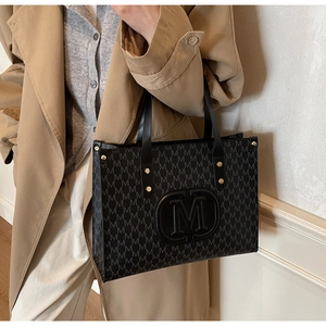 Close-up of a black tote bag with M-pattern print worn with a khaki trench coat