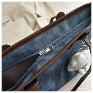 Close-up of a blue denim tote bag with brown leather trim, silver zipper, and plush charm