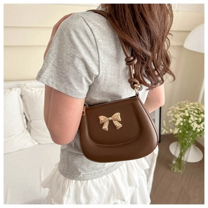 Brown faux leather saddle bag with gold bow buckle worn by a model in a cozy interior