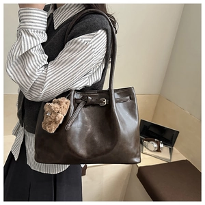 Detail of a dark brown leather tote bag with a buckle strap and plush bear charm