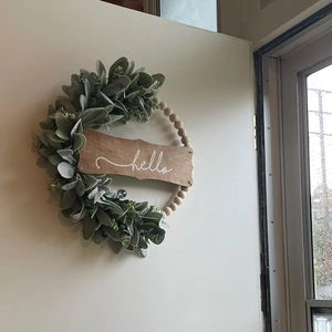 Detail of wooden bead wreath with sage green faux leaves and hello sign on white wall