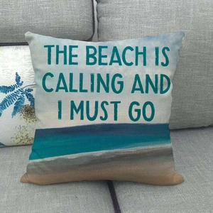 Linen cushion cover with beach scenery and 'The Beach is Calling' text on a grey sofa