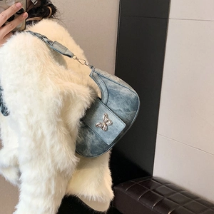 Blue denim half-moon underarm bag with silver butterfly buckle shown on white coat