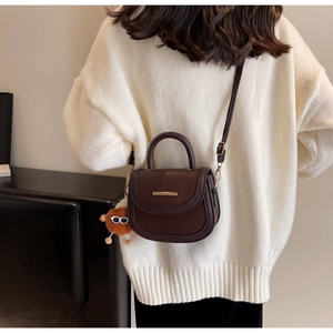 Détail du sac selle marron foncé avec garniture dorée et breloque peluche sur pull blanc