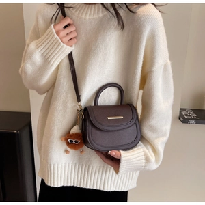 Détail d'un sac besace marron foncé avec barre dorée et breloque peluche sur pull blanc
